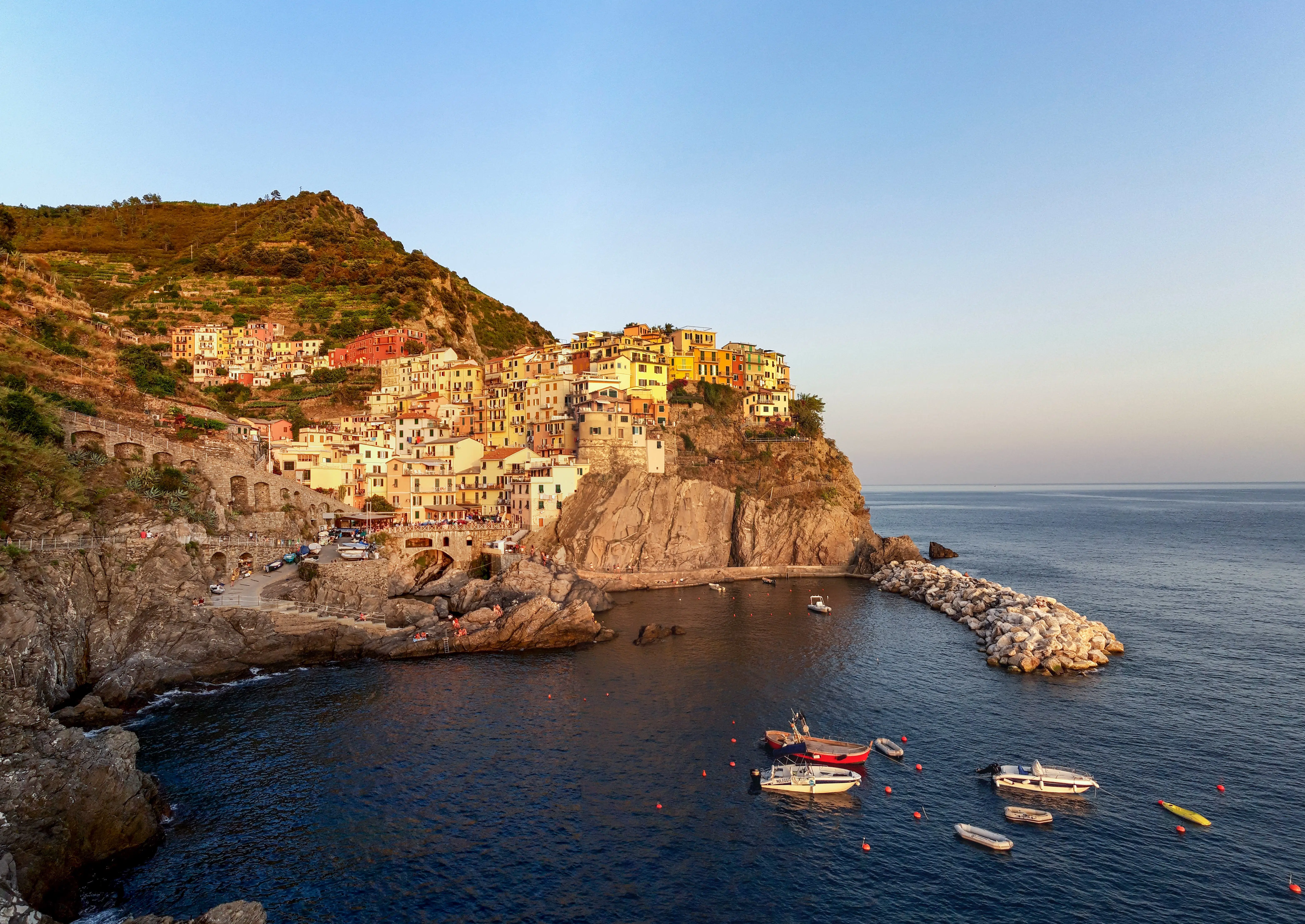 Prédios e casas coloridas amontoadas na costa elevada de Cinque Terre, em Manarola, na Itália, perto do mar onde barcos passeiam. A imagem ilustra o guia sobre quanto custa um seguro viagem Europa. - Foto: Matej Drha via Unsplash