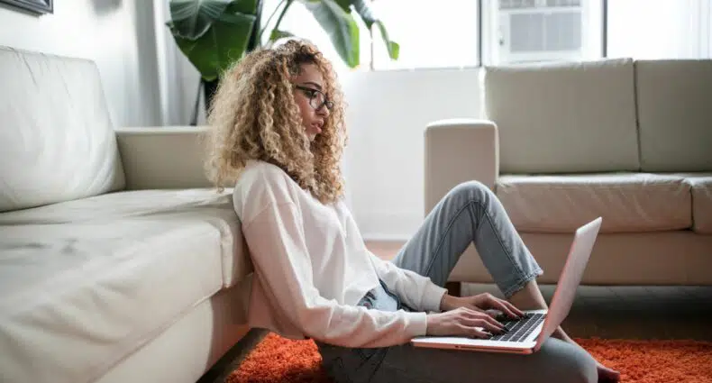 Mulher usando computador, sentada no chão e recostada no sofá