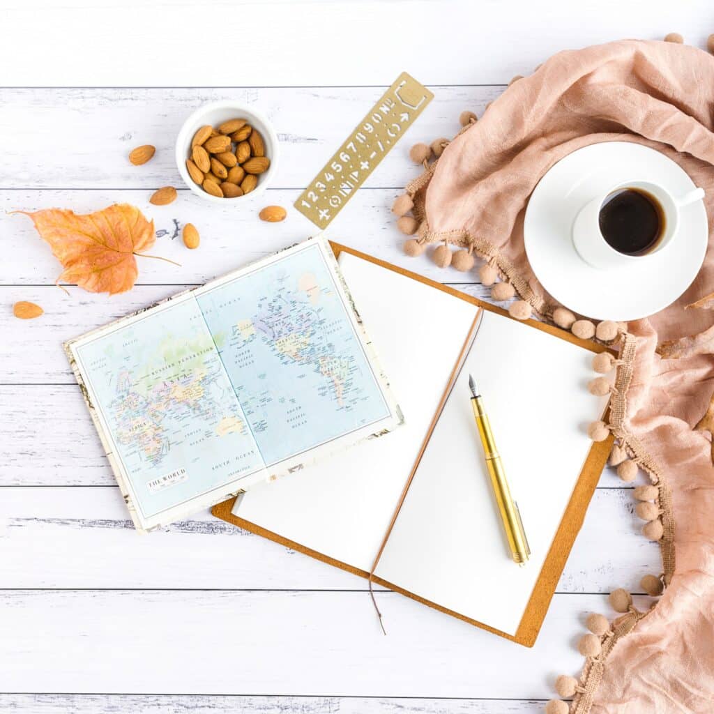 caderneta aberta com folhas em branco, uma caneta em cima, uma xícara de café ao lado e um mapa mundi aberto