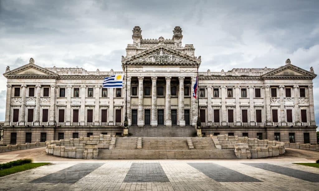 Palácio Legislativo de Montevidéu. O local é da cor branca evelhecida e tem várias colunas e janelas. Uma bandeira do Uruguai está tremulando ao vento logo em frente.