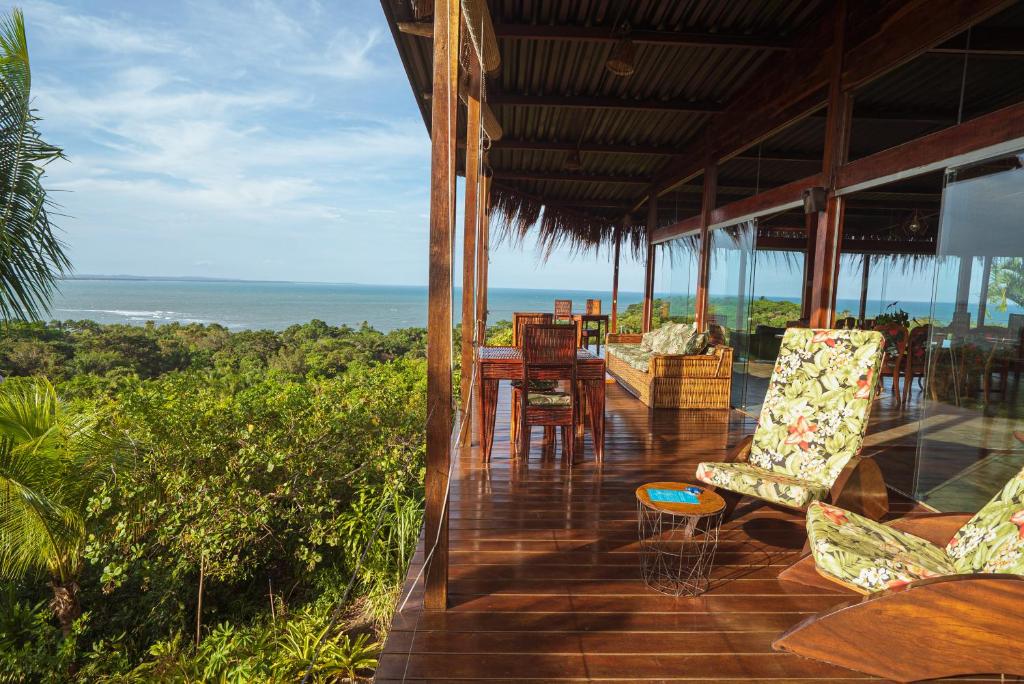 Um deck de madeira da Pousada O Céu de Boipeba com cadeiras e mesas tem vista para uma vegetação exuberante e para o oceano sob um céu parcialmente nublado.