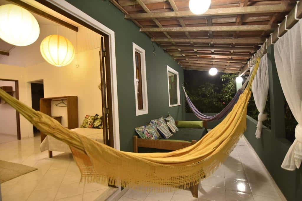 Pátio coberto da Pousada Sossego com paredes verdes, duas redes, banco almofadado e vista para uma sala interna com iluminação aconchegante.