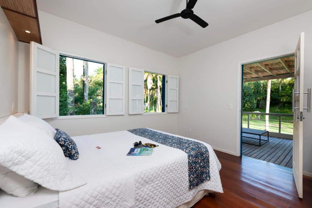 Um quarto da Pousada Vila da Barra com uma cama, paredes brancas, duas janelas abertas, um ventilador de teto e uma porta que dá para uma varanda com vista para as árvores.