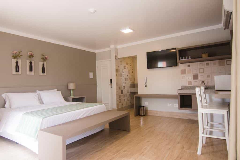 Um quarto na Pousada Villa Di Enzo no Itaguá com uma cama de casal, escrivaninha, TV, bancada e uma pequena área de cozinha com decoração em cores claras.
