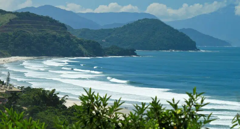 Paisagem costeira com uma praia de areia, vegetação verdejante em primeiro plano e colinas ondulantes sob um céu azul claro.
