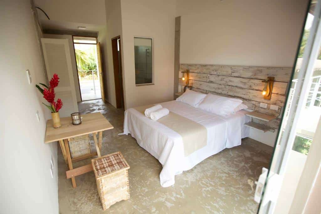 Um quarto na Vila Sapê Pousada com uma cama de casal, detalhes em bege, uma pequena mesa de madeira e duas luminárias de parede, com uma porta para o exterior, para representar onde ficar em Ubatuba.