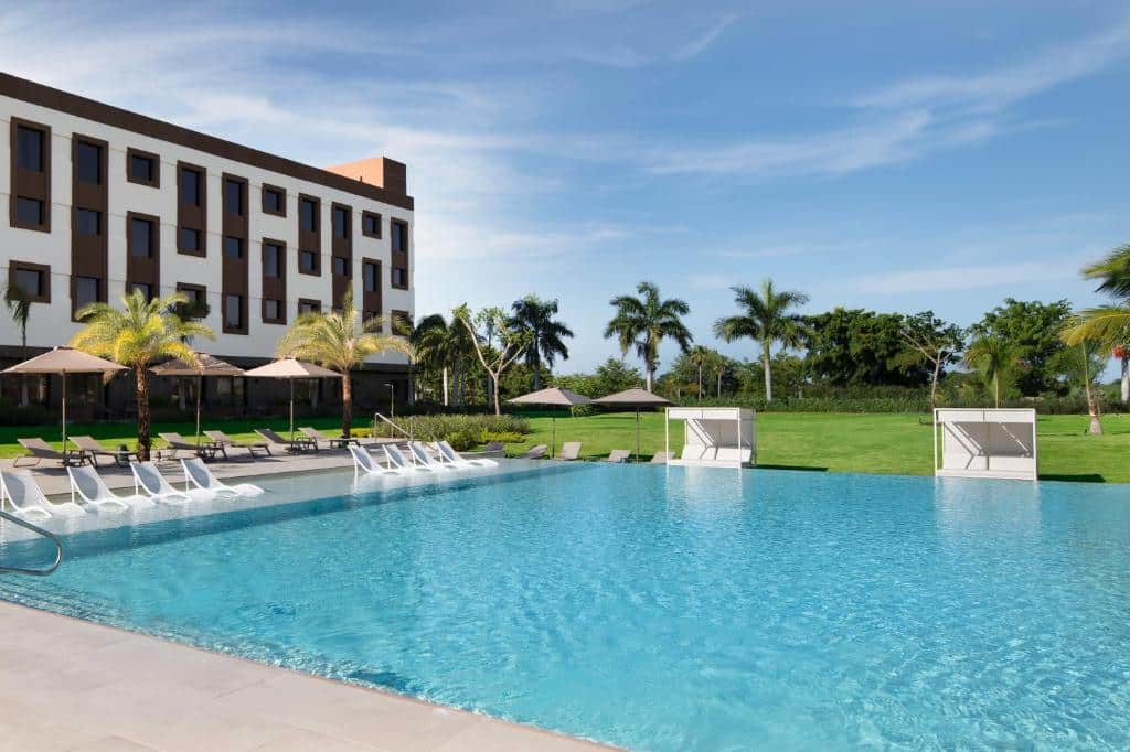 Piscina do AC Hotel by Marriott Punta Cana rodeada por espreguiçadeiras e guarda-sóis. Há um gramado ao fundo, e no canto esquerdo fica o prédio do hotel.