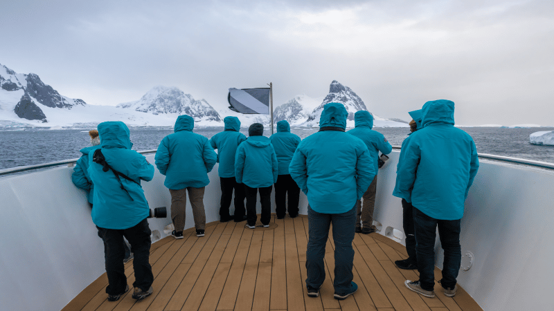 Pessoas de agasalho azul dentro de um barco. Elas estão de costas para a foto e olham para os montes cobertos de gelo na Antártica.