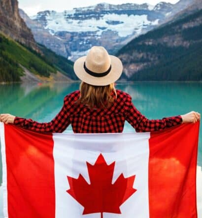 Mulher vestindo camisa xadrez vermelha e preta e chapéu bege segurando a bandeira do Canadá nas costas enquanto olha para um lago à sua frente. Há montanhas desfocadas ao fundo. - Foto: Andre Furtado
