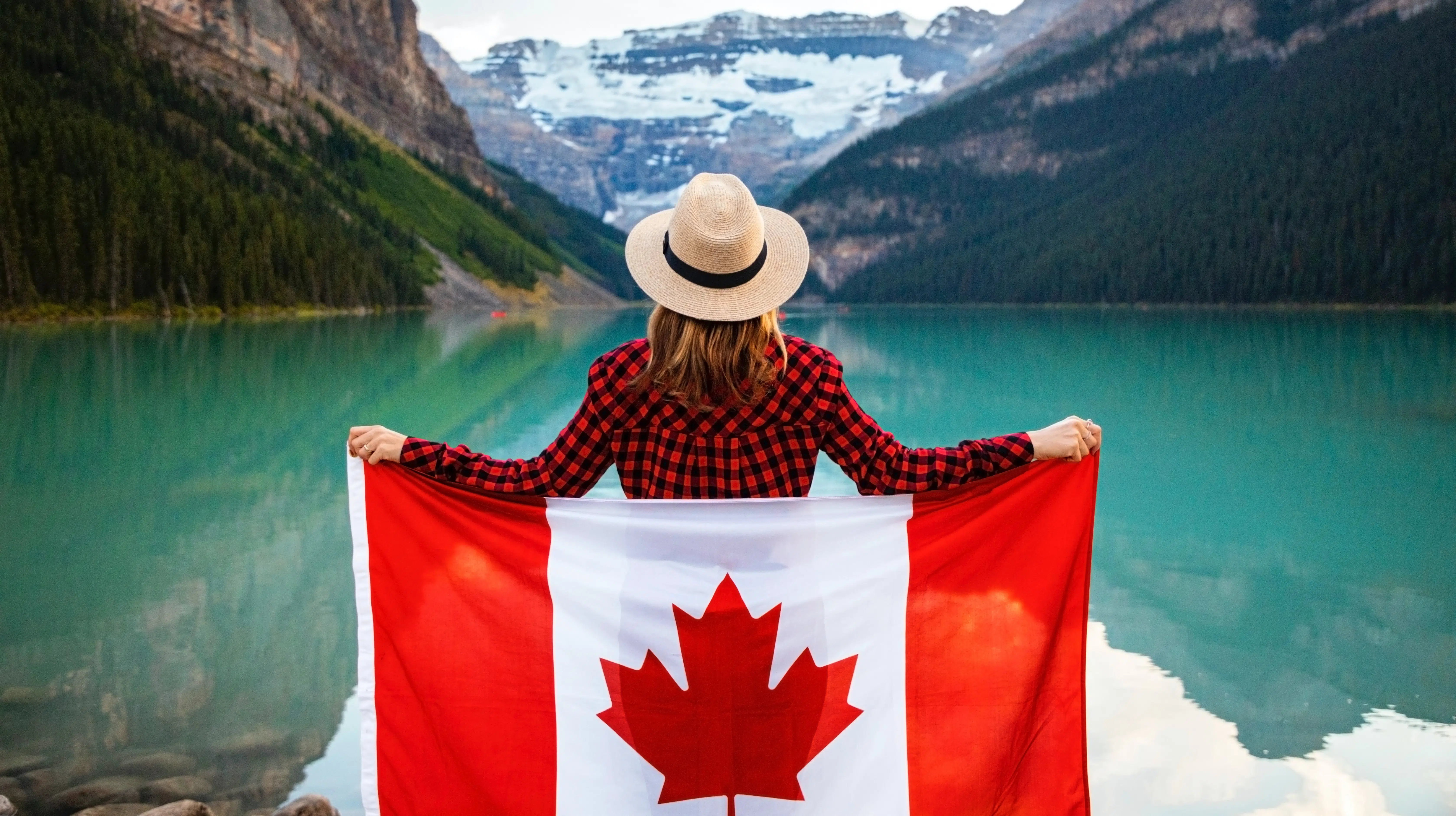 Mulher vestindo camisa xadrez vermelha e preta e chapéu bege segurando a bandeira do Canadá nas costas enquanto olha para um lago à sua frente. Há montanhas desfocadas ao fundo. - Foto: Andre Furtado