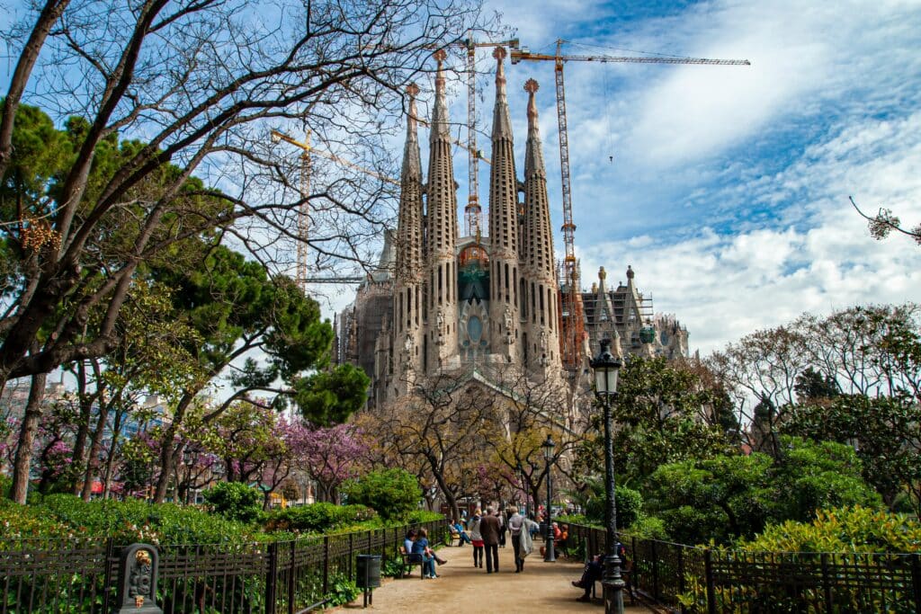 entrada catedral sagrada família que pode ser visitada com um seguro viagem Barcelona. o local é cercado por árvores, e no centro há uma passagem onde várias pessoas passeiam.