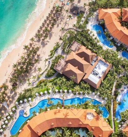 Vista aérea do Majestic Elegance Punta Cana Resort, um dos resorts em Punta Cana. Piscinas estão espalhadas entre os prédios do local, e há várias árvores ao redor. Do lado esquerdo da imagem está a praia.