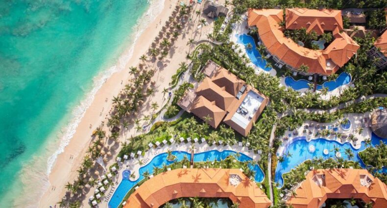 Vista aérea do Majestic Elegance Punta Cana Resort, um dos resorts em Punta Cana. Piscinas estão espalhadas entre os prédios do local, e há várias árvores ao redor. Do lado esquerdo da imagem está a praia.