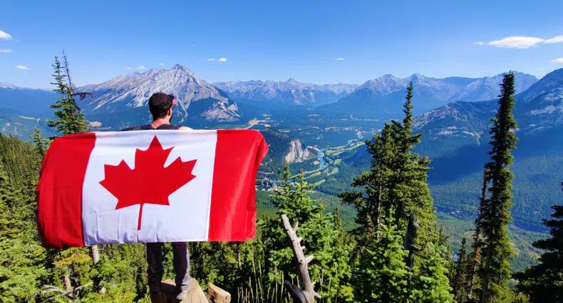 Pessoa em pé no penhasco de uma montanha, segurando uma bandeira canadense, com vista para um vale com montanhas e árvores sob um céu azul claro. A imagem ilustra o post sobre intercâmbio no Canadá.