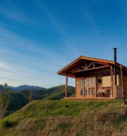 Cabana de madeira do Chalé na Pousada Capim Limão, ficando no topo de uma colina gramada com montanhas e um céu azul ao fundo.
