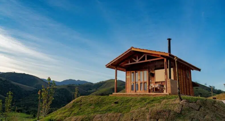 Cabana de madeira do Chalé na Pousada Capim Limão, ficando no topo de uma colina gramada com montanhas e um céu azul ao fundo.