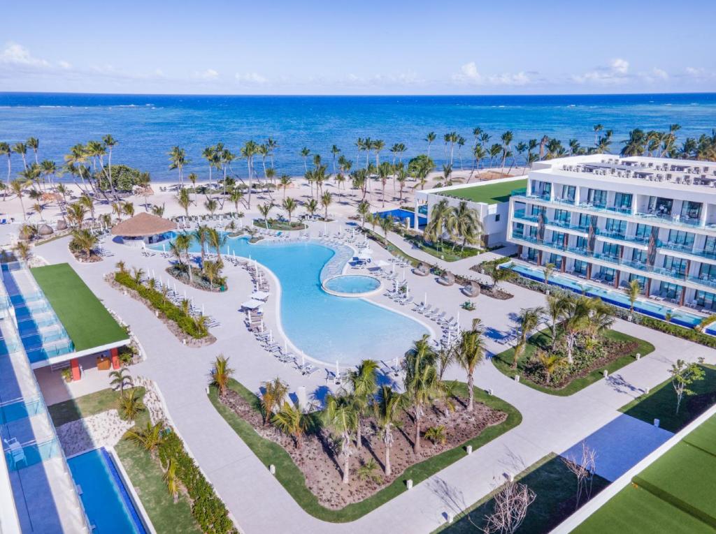 Vista aérea do Serenade Punta Cana Beach & Spa Resort. Uma piscina ampla fica no centro do local e é rodeada por espreguiçadeiras e áreas verdes com palmeiras. Os prédios do resort ficam nas laterais, e é possível ver a praia ao fundo.