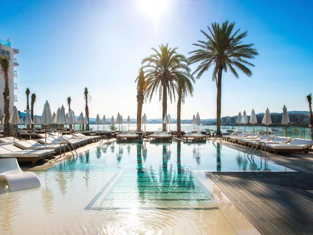 Piscina do Amàre Beach Hotel Ibiza, com águas cristalinas refletindo o céu e algumas árvores, céu azulado com um raio de sol branco no meio e várias poltronas brancas em volta com guarda-sóis fechados