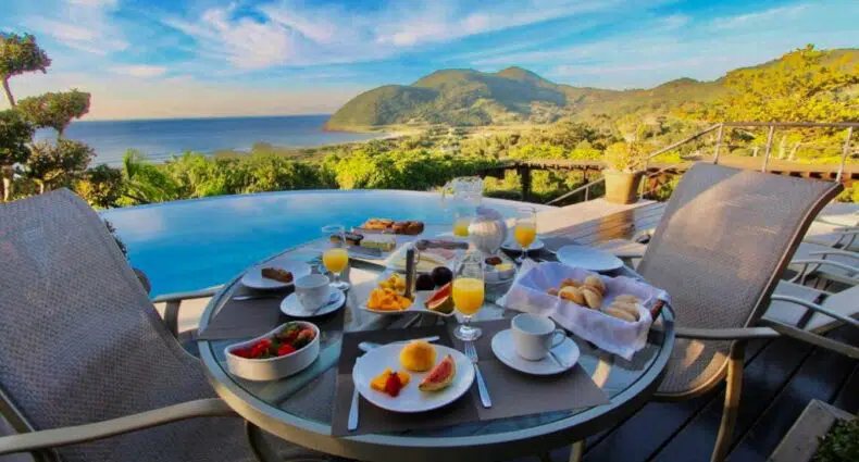 Uma mesa de vidro posta para com o Café da manhã da Morada Prainha, uma das pousadas em Garopaba. Há vista para uma piscina, montanhas e o oceano sob um céu azul com nuvens.