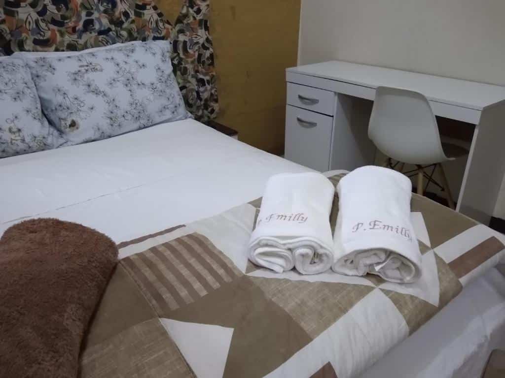 cama de casal com duas toalhas e dois acolchoados na Pousada Emilly em Joinville. No lado direito da imagem é possível ver uma mesa branca com cadeira.