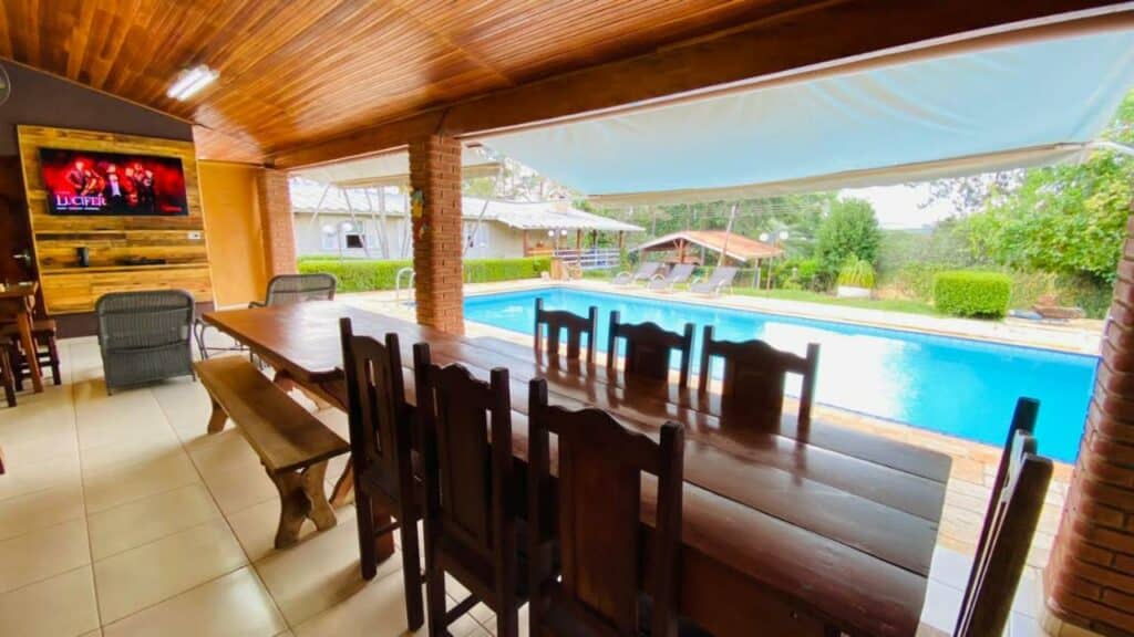 Área externa com piscina no Chalé , piscina, churrasqueira com mesa com sete cadeiras a frente e ao fundo a piscina.
