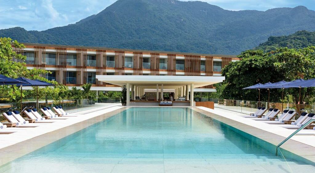 Piscina externa retangular com espreguiçadeiras ao lado de um moderno edifício de hotel e montanhas ao fundo, no Hotel Fasano Angra dos Reis.