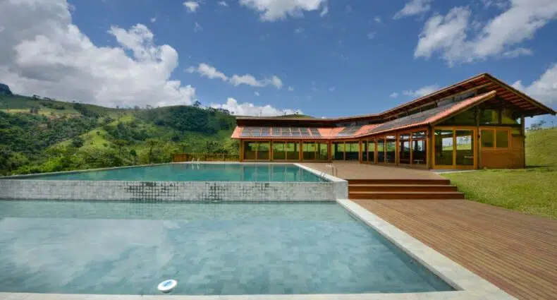 Casa moderna com grandes janelas e um deck de madeira ao lado de uma piscina retangular, situada em uma paisagem verde e montanhosa, na Pousada Quinta de Santa Bárbara, para representar pousadas em São Francisco Xavier