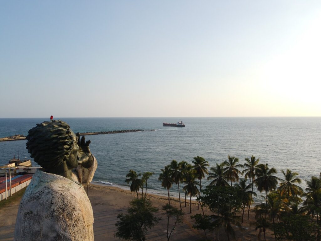 Monumento de Fray Anton de Montesinos, estátua em pedra de um homem olhando para o mar, próximo de uma praia com alguns coqueiros, para representar o seguro viagem Santo Domingo