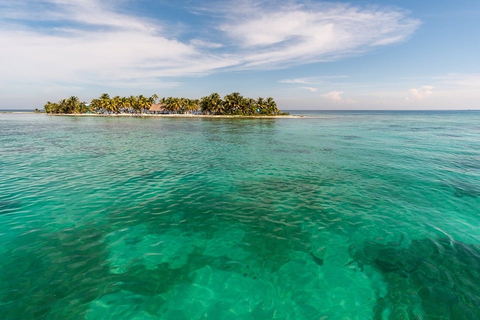 Mar de águas azuis cristalinas e a ilha de Caye ao fundo com muitos coqueiros sob um céu azul ensolarado. Seguro Viagem Belize.