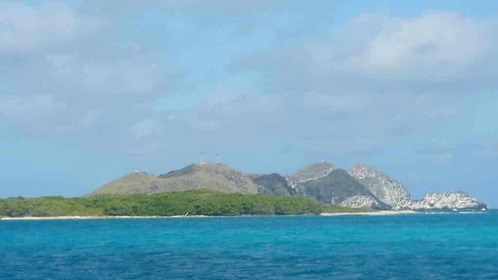 Ilha Madrisquí em Los Roques formado por um complexo de pedras ao fundo, áreas verdes e um mar de tom azul cristal em um dia ensolarado e céu azul.