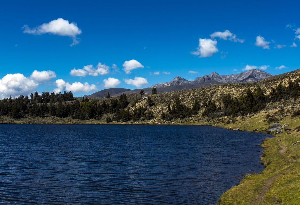 Lago glacial no Parque Nacional de Sierra Nevado com uma área verde ao redor e montanhas ao fundo em um dia de céu azul e ensolarado.
