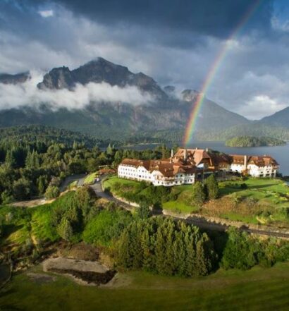 Hotel em Bariloche com vista para o lago e montanhas, arco-íris no céu nublado