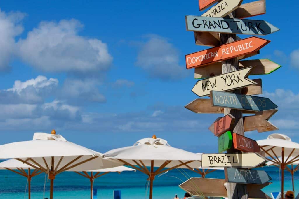 Grace Bay, praia com mar de azul escuro, muitos guarda-sóis brancos e, em destaque na foto, um poste com diversas placas coloridas com nomes de países indicando direções diferentes
