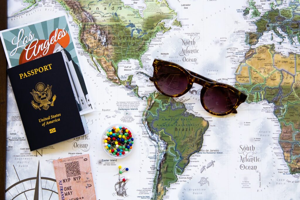 Foto de mapa com passaporte americano, print escrito "Los Angeles", um óculos de sol, uma caixinha com alfinetes e uma passagem aérea