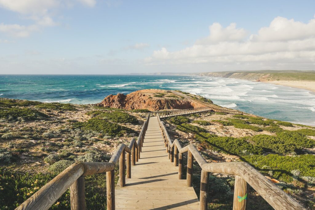 Caminho com corrimão de madeira, por entre pedras e vegetação, na Praia da Bordeira, em direção ao mar