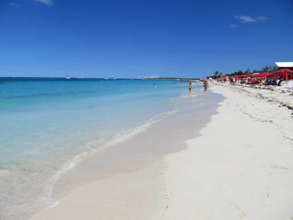 Praia Orient Bay em St Martin, uma praia de areia extremamente branca e mar tão azul que fica transparente, é possível ver guarda-sóis vermelhos presos na areia e algumas pessoas caminhando