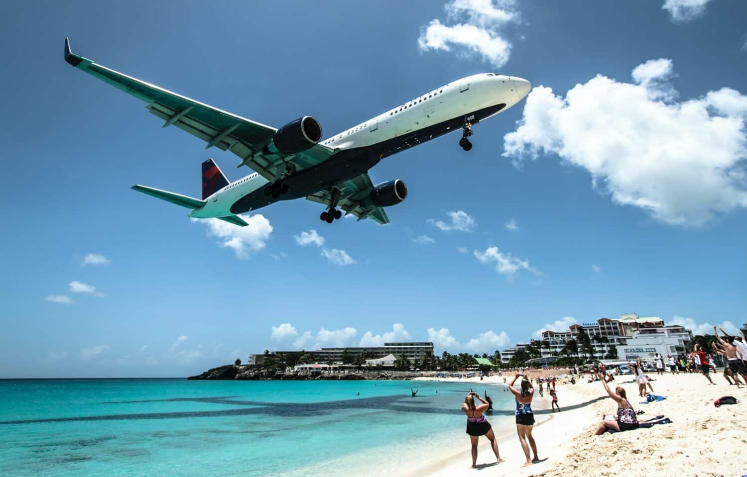 Um avião sobrevoando a praia Maho em St Maarten, o mar é de um verde claro, a areia é branca e alguns turistas estão parados fotografando o avião