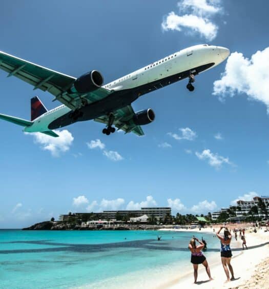 Um avião sobrevoando a praia Maho em St Maarten, o mar é de um verde claro, a areia é branca e alguns turistas estão parados fotografando o avião