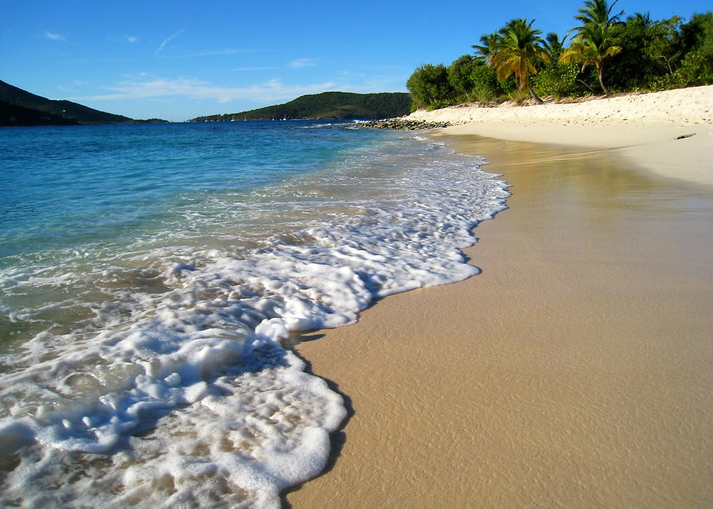 Sandy Cay, uma praia com uma certa elevação na parte da areia, dividindo ela em uma parte mais dura e outra mais macia, o mar é azul claro e, ao fundo, é possível ver vegetação após a faixa de areia