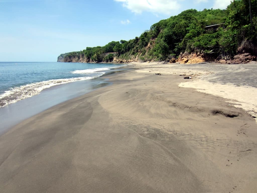 Praia Woodlands com a areia mais escura por conta das erupções vulcânicas na ilha, o mar é azul, e próximo da areia tem rochas e vegetação
