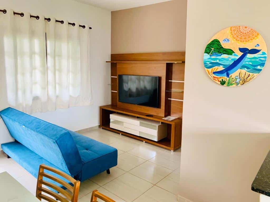 Sala de estar em Apto 2 Qtos c/ Ar Cond 400Mt Praia do Centro, uma pequeno sofá, um rack com televisão, uma janela com cortina e um quadro colorido