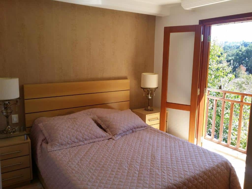 Quarto no Aptos de 03 e 04 quartos Centrinho Capivari, uma cama de casal, dois abajures, uma sacada com vista para as árvores, para representar airbnb em Campos do Jordão