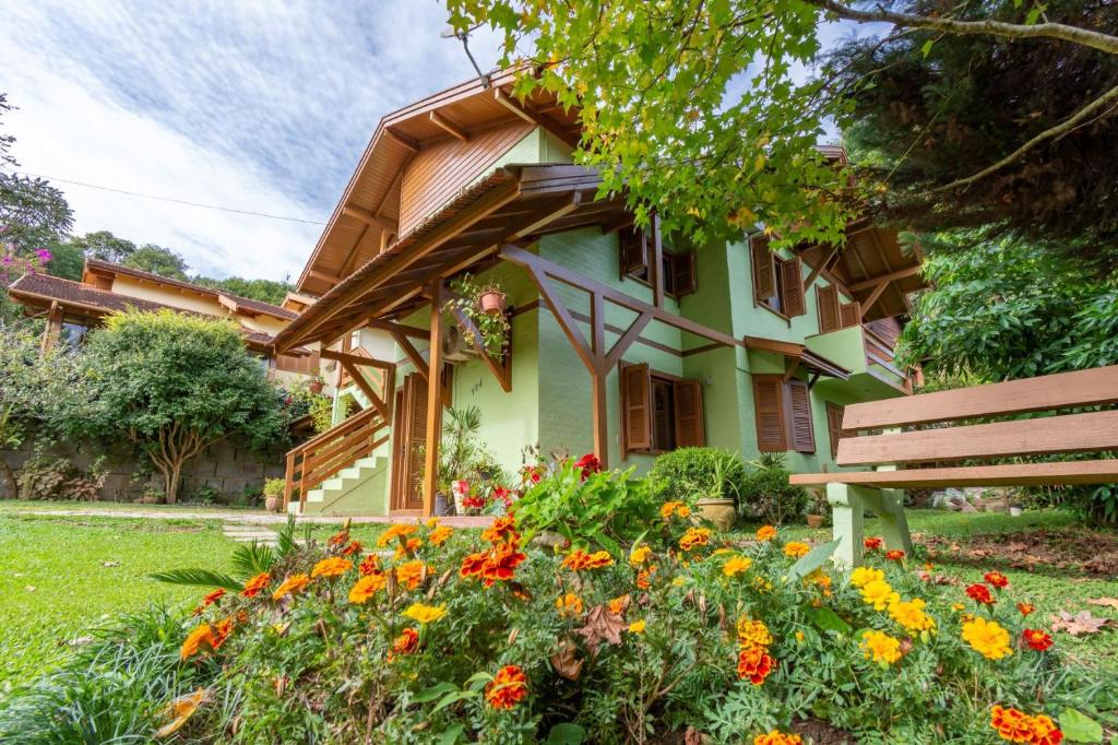 Entrada da Rosa Casa Tirol Gramado, cercada por um gramado com flores e árvores, a construção tem dois andares, é pintada em um tom de verde claro com detalhes em madeira