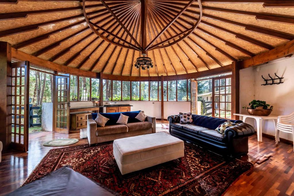 Sala da Casa Xamego Alto Padrão, local amplo, inteiro com janelas e portas de vidro virados para a mata, dois sofás, uma tapete, estrutura do teto em madeira, para representar airbnb em Campos do Jordão