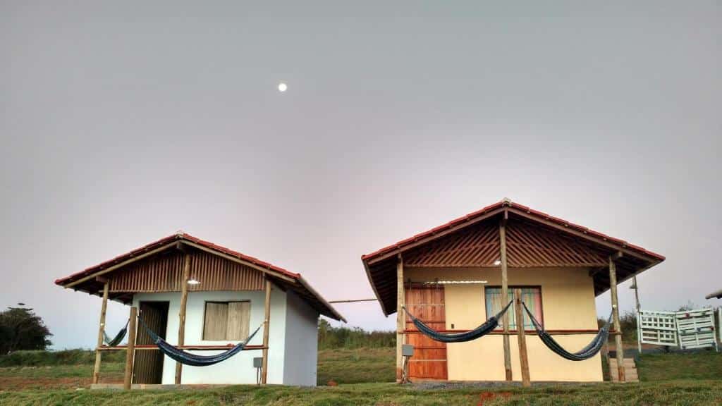 Dois chalés da Aldeia Canastra Pousada com varanda e redes sob um céu nublado com a lua ao fundo. Cada chalé é de uma cor diferente. À esquerda um chalé verde e à direita um chalé amarelo