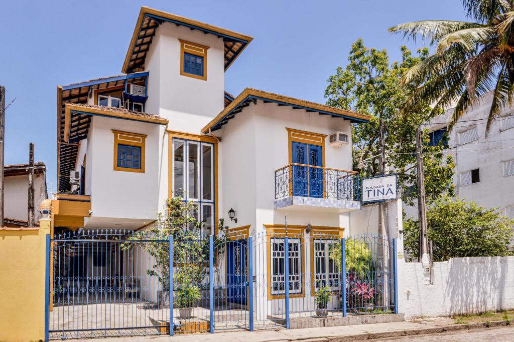 Prédio da Pousada da Tina, parece com uma casa antiga, inteira pintada de branco com detalhes charmosos em azul e amarelo, um portão baixo e muitas janelas