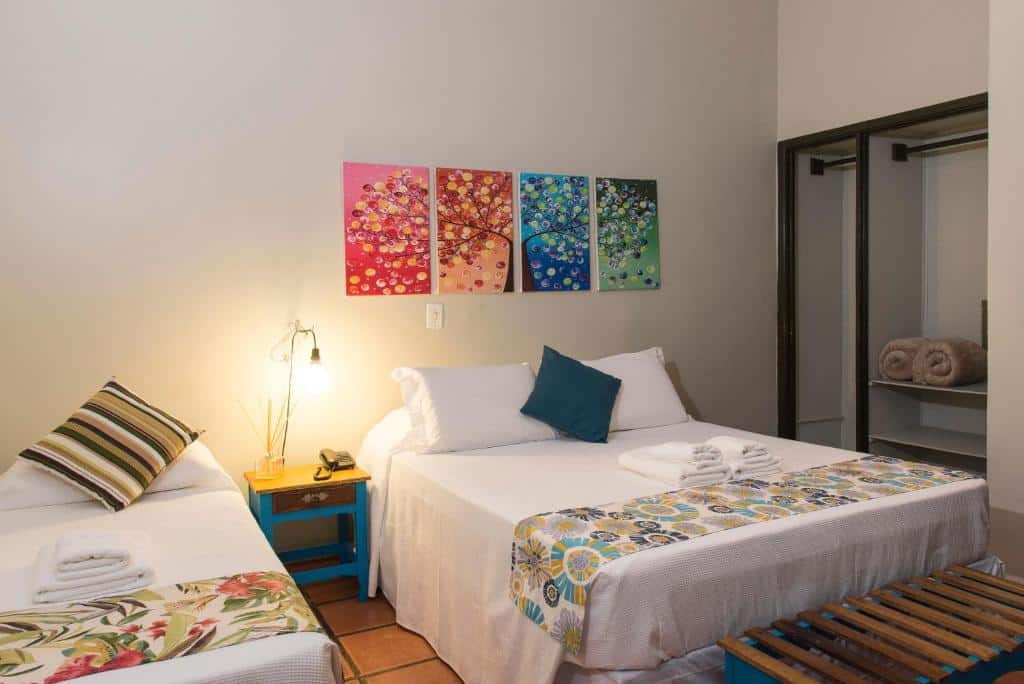 Quarto no Hotel Porto Di Mare, uma cama de casal, uma de solteiro, uma mesinha com um abajur, quadros coloridos, um armário de conceito aberto, almofadas e travesseiros sob a cama, para representar airbnb na Praia do Lázaro