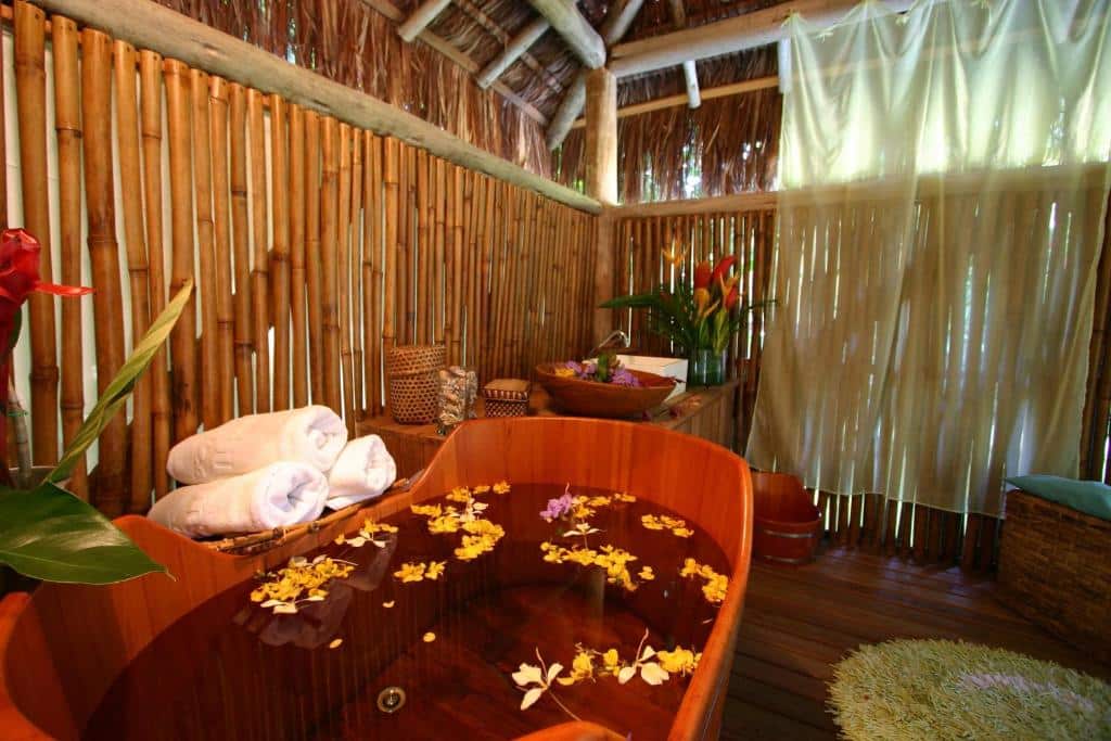 Banheira cheia de água e flores no Clique na imagem e faça sua reserva no Itamambuca Eco Resort. Ela está em uma sala com paredes de bambu e com vários utensílios para serviço de spa.