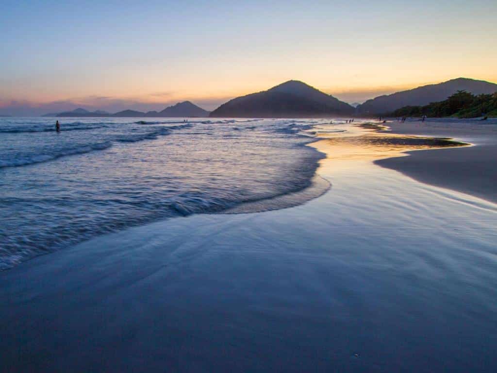 Praia da Itamambuca no fim da tarde, extensa faixa de areia, mar calmo e vista para as montanhas