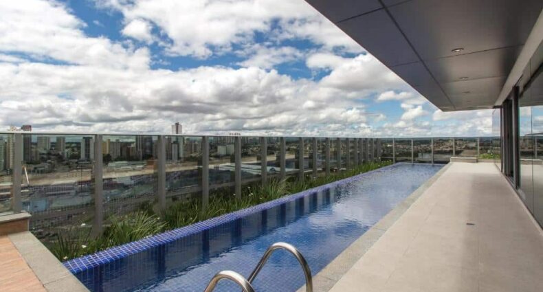 Uma piscina estreita na cobertura, com guarda-corpo de vidro, tem vista para uma paisagem urbana sob um céu parcialmente nublado.
